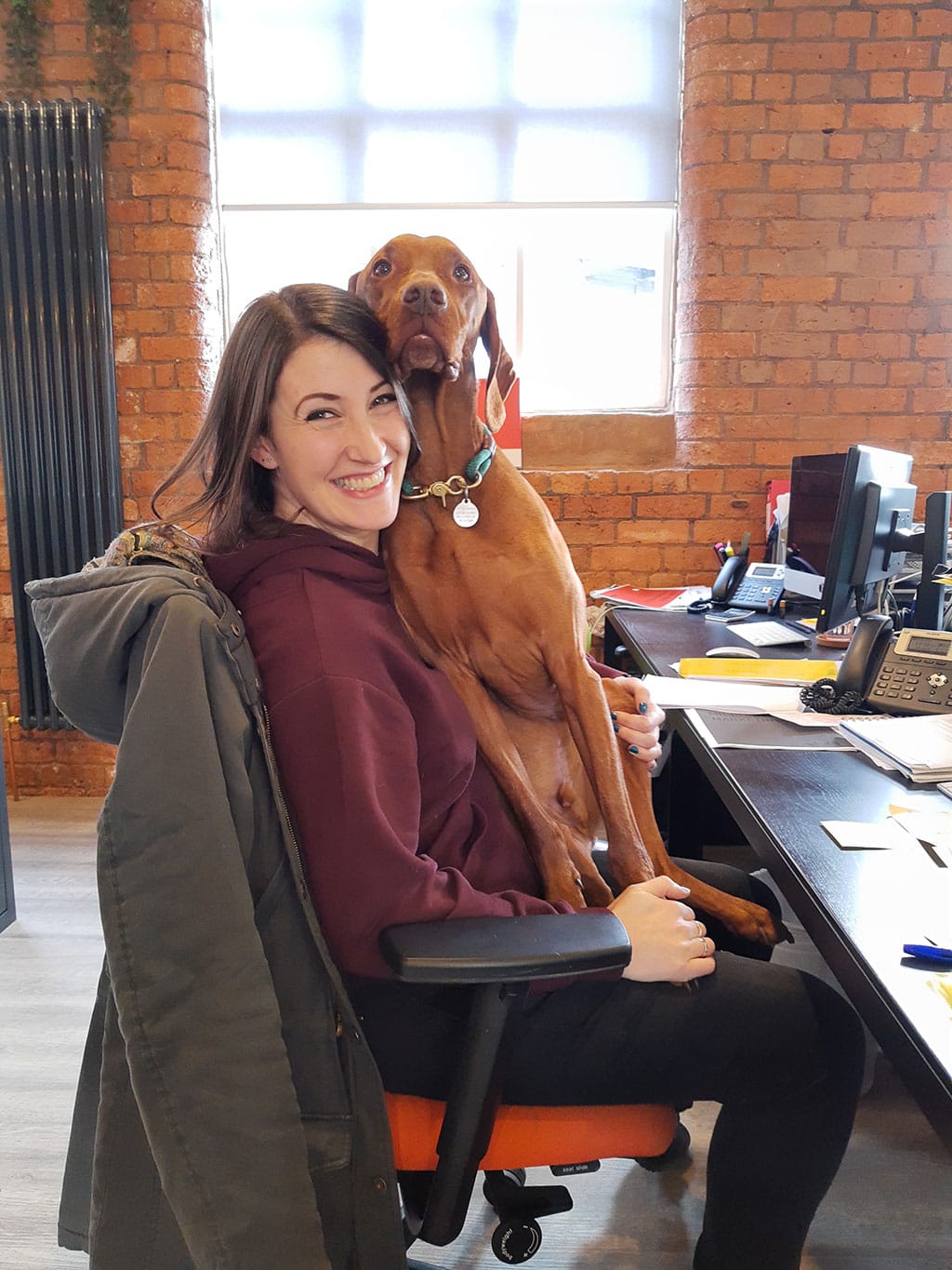 A photo of Vivid's Christina with her dog, Rory, on her lap, sat at their desk smiling to the camera