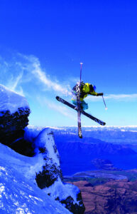 Skier performing a high aerial jump against a vivid blue alpine sky in snowy mountain scenery.