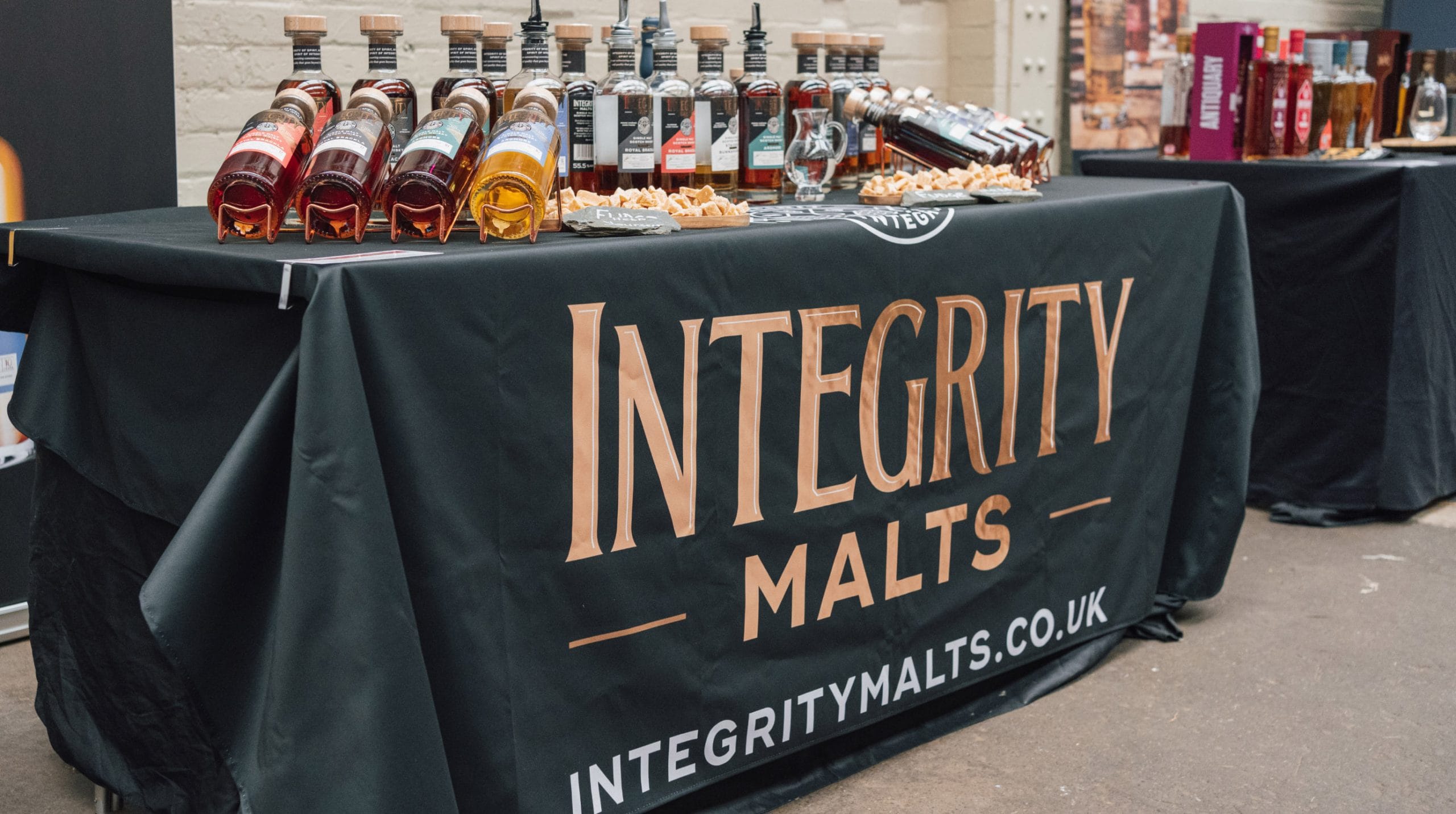 Integrity Malts branded exhibition stand displaying whisky bottles, tasting equipment and promotional table covering at a trade show or tasting event.